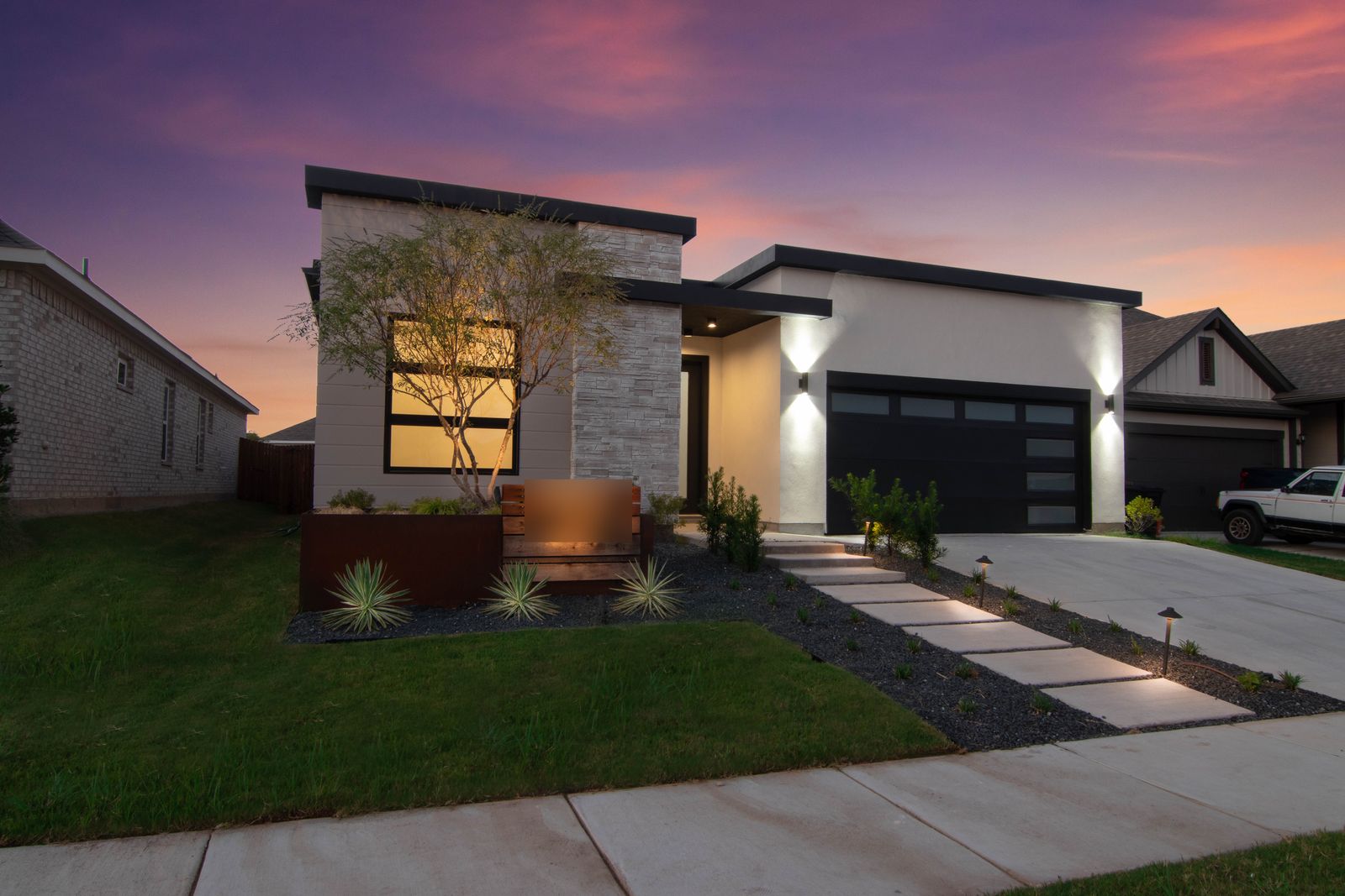 Modern stone-front home at dusk with warm window light and a landscaped yard